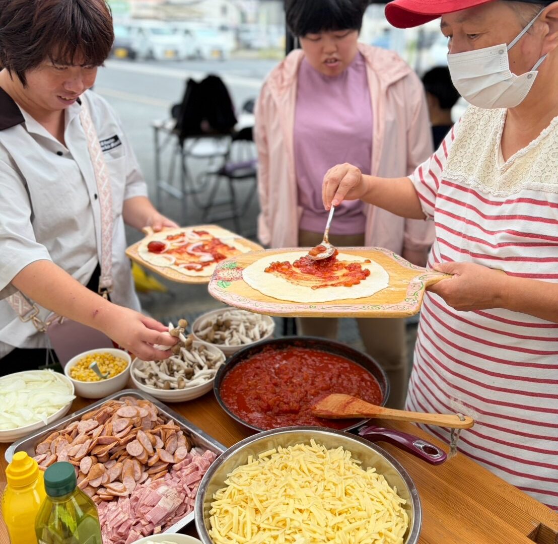 チーズや野菜などの具材が並ぶテーブルでピザをつくる様子