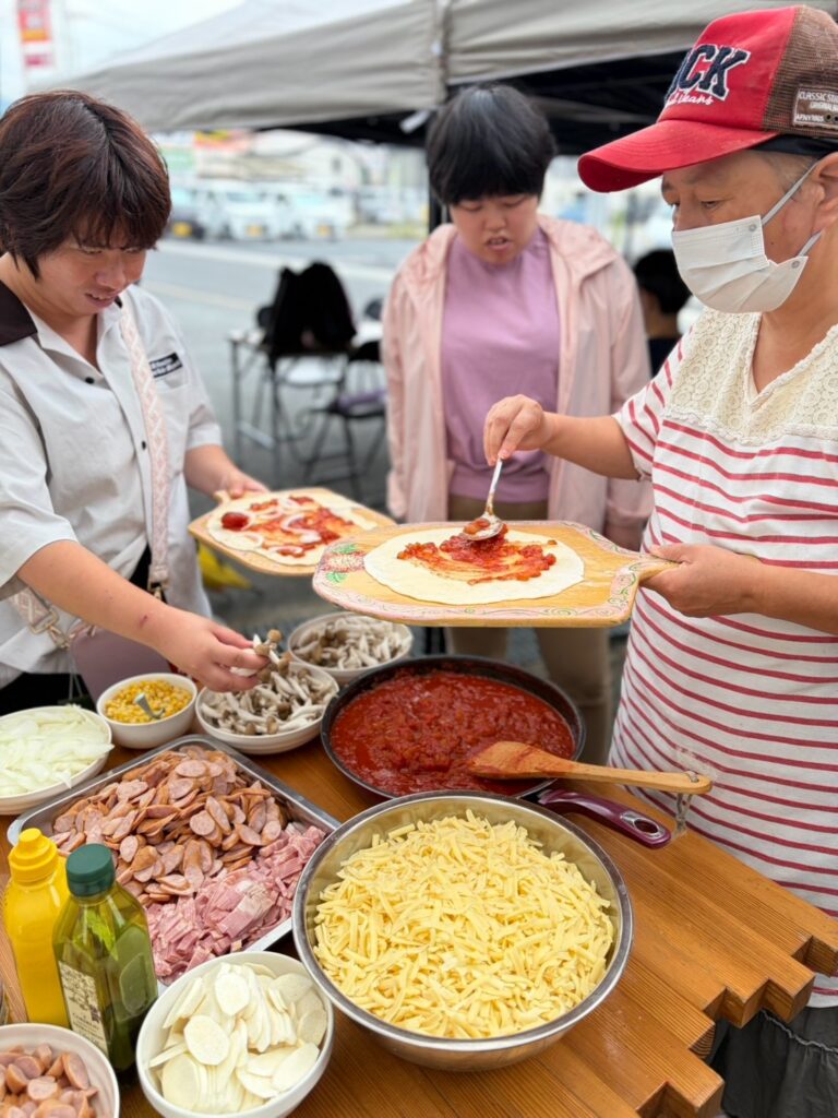チーズや野菜などの具材が並ぶテーブルでピザをつくる様子